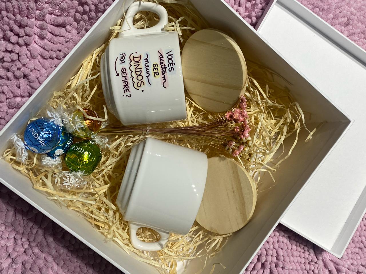 Caixa presente com caneca e copos de madeira, bombons coloridos e confetes dourados sobre fundo roxo.